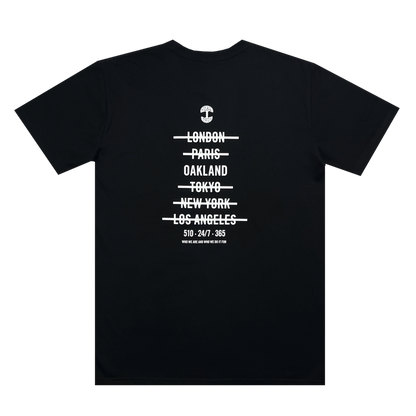 The Oaklandish "Who We Are and Who We Do it For Tee" is a 100% cotton, classic fit T-shirt with white text in a minimalist design. The text features a small anchor graphic at the top, followed by city names with lines through them: "London, Paris, Oakland, Tokyo, New York, Los Angeles." Below the cities is the text: "510-247-365".
