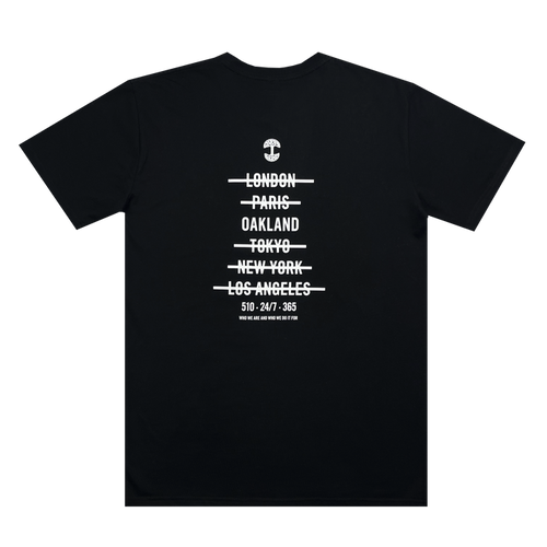 The Oaklandish "Who We Are and Who We Do it For Tee" is a 100% cotton, classic fit T-shirt with white text in a minimalist design. The text features a small anchor graphic at the top, followed by city names with lines through them: "London, Paris, Oakland, Tokyo, New York, Los Angeles." Below the cities is the text: "510-247-365".