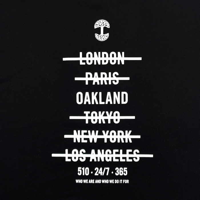 The "Who We Are and Who We Do it For Tee" by Oaklandish features a black background with white text. At the top is a tree logo. Below the logo, the names of cities—"London," "Paris," "Oakland," "Tokyo," "New York," and "Los Angeles"—are written and crossed out. Underneath these, on a classic fit t-shirt made of 100% cotton, are the phrases: "510 - 24/7 - 365" and "Who we are and who we do it for.