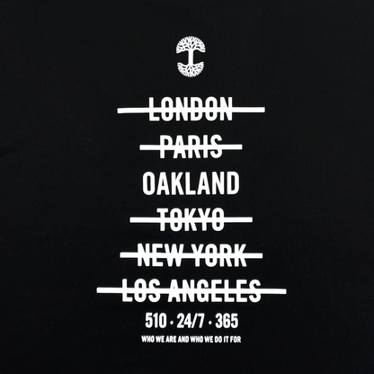 The "Who We Are and Who We Do it For Tee" by Oaklandish features a black background with white text. At the top is a tree logo. Below the logo, the names of cities—"London," "Paris," "Oakland," "Tokyo," "New York," and "Los Angeles"—are written and crossed out. Underneath these, on a classic fit t-shirt made of 100% cotton, are the phrases: "510 - 24/7 - 365" and "Who we are and who we do it for.