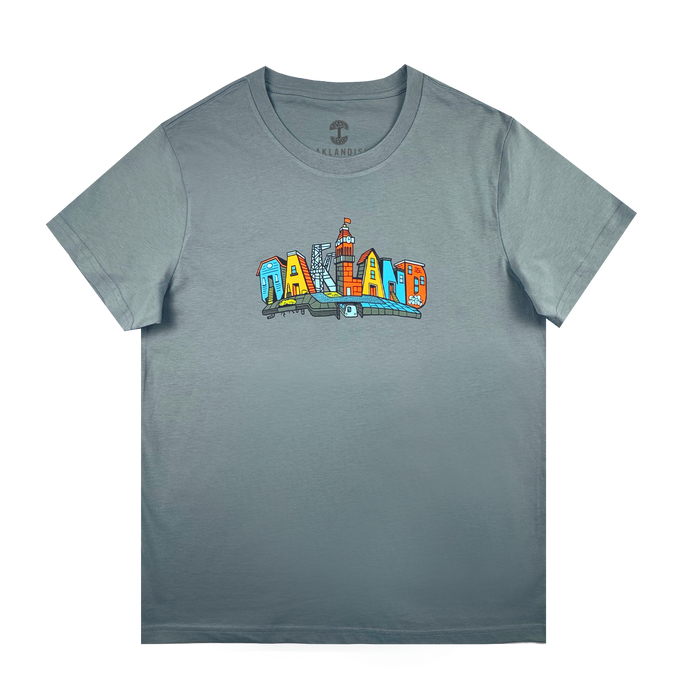 The Oaklandish Women's Blockletters Tee is a gray cotton shirt with "OAKLAND" in bold, colorful cartoon-style letters featuring city elements and skyline details, all above a subtle shadow graphic.