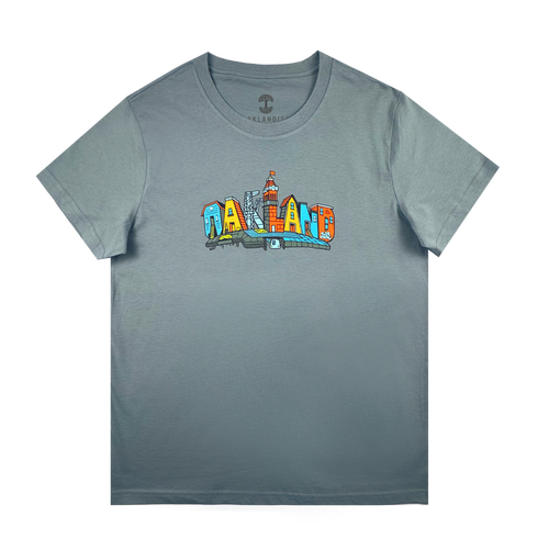 The Oaklandish Women's Blockletters Tee is a gray cotton shirt with "OAKLAND" in bold, colorful cartoon-style letters featuring city elements and skyline details, all above a subtle shadow graphic.