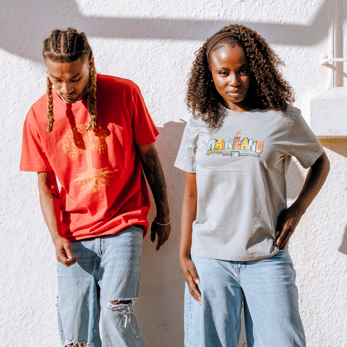 Two people stand against a sunlit white wall. The left wears a red Pemex shirt and light jeans; the right sports an Oaklandish Women’s Blockletters Tee in light gray with matching jeans. Both have curly hair and look relaxed.