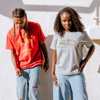 Two people stand against a sunlit white wall. The left wears a red Pemex shirt and light jeans; the right sports an Oaklandish Women’s Blockletters Tee in light gray with matching jeans. Both have curly hair and look relaxed.